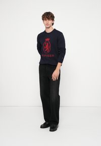 Maglione blu navy con un emblema di un leone rosso, abbinato a jeans neri e scarpe nere. Il maglione presenta una scollatura tonda e polsini a coste.