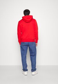 Sudadera con capucha roja hecha de tela suave, combinada con jeans anchos azules y zapatillas blancas. La sudadera tiene un bolsillo canguro y puños acanalados.