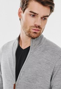 Szary sweter na zamek błyskawiczny z wysokim kołnierzem, o delikatnej fakturze. Noszony na czarnej koszulce z dekoltem w serek, z metalowym zamkiem błyskawicznym i skórzaną zawieszką.