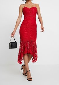 Robe en dentelle rouge avec un décolleté en cœur et un corsage ajusté. Possède un ourlet asymétrique et des détails en dentelle transparente. Sac à main noir et talons à lanières.
