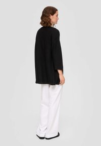 Schwarzer oversized Pullover mit lockerer Passform, breiten Ärmeln und seitlichen Schlitzen. Kombiniert mit weiten weißen Hosen und schwarzen flachen Schuhen.