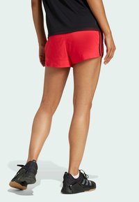 Shorts de sport rouges en tissu lisse, avec des rayures noires sur les côtés, associés à des baskets noires et un t-shirt noir.