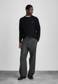 Μαύρο ριμπ μπουφάν με στρογγυλή λαιμόκοψη, συνδυασμένο με γκρι πλισέ παντελόνι και μαύρα παπούτσια, σε ένα μίνιμαλ φόντο.