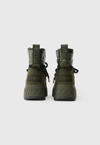 Olive spalvos kulkšninių batų su siuvinėta viršutine dalimi, tekstūruotais gumos padu, prekės ženklo akcentais ir juodais raišteliais, sukurti komfortui ir stiliui.