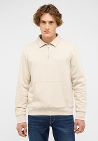 Beżowa bluza z zamkiem na kołnierzu, kieszenią kangurką i subtelnym haftem z okrągłym logo. Miękki materiał, luźny krój i ściągacze na rękawach.
