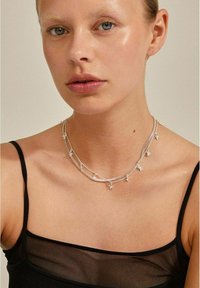 Sølv halskæde med flere lag af kæder, der har små blomsterdetaljer. Modellen bærer en sort gennemsigtig top, der tydeligt fremhæver halskæden.