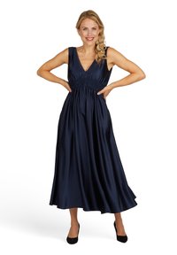 Robe satin bleu marine avec un décolleté en V et des bretelles larges, mettant en valeur un corsage smocké et une jupe fluide longue jusqu'aux chevilles. Des talons noirs complètent le look.