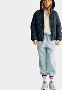 Veste matelassée bleu marine avec capuche, pantalon de survêtement gris clair et chaussettes rayées multicolores. Des baskets avec des accents réfléchissants complètent le look.