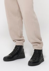 Czarne sneakersy za kostkę z teksturowanej skóry, okrągłym noskiem i sznurowanym zapięciem, noszone z beżowymi spodniami dresowymi z podwiniętymi nogawkami.