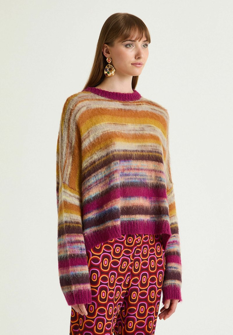 Donna che indossa un maglione di pelliccia colorato a righe e pantaloni in stile retrò con motivi, accessoriata con grandi orecchini pendenti multicolori.