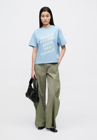 Ανοιχτό μπλε βαμβακερό T-shirt με έντονο λευκό κείμενο. Φόρμες cargo σε χακί χαλαρή γραμμή. Μαύρη τσάντα με υφή και μυτερές μαύρες παπούτσια.