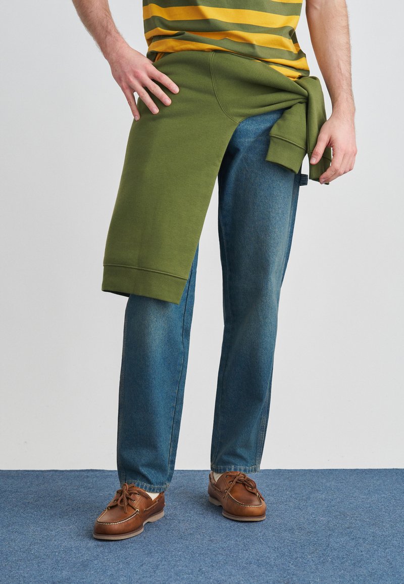 Indumento in stile felpa verde drappeggiato su jeans blu. I jeans hanno un taglio dritto, mentre la felpa presenta polsini a coste e una vestibilità ampia.