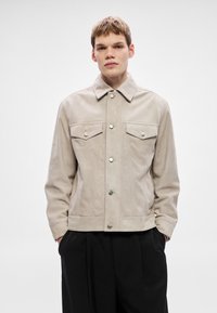 Bézs színű, szarvasbőr dzseki gombos elülső résszel, hegyes gallérral és két mellzsebbel. Ezüst színű gombokkal és testhezálló fazonnal rendelkezik.