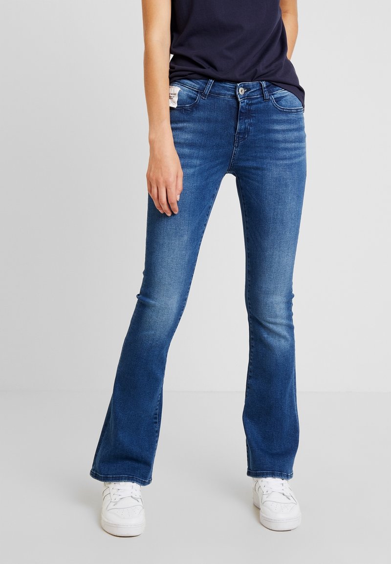 Personne portant un jean bootcut bleu moyen, des baskets blanches et un haut bleu marine foncé, debout devant un fond clair uni.