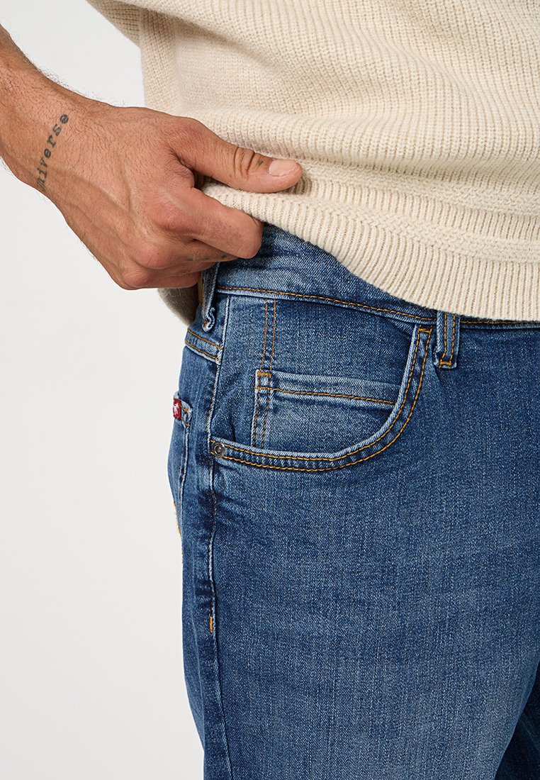 Niebieskie dżinsy o dopasowanym kroju, z detalicznym przeszyciem, pięcioma kieszeniami i małą metką markową na tylnej kieszeni. Tekstura jest gładka.