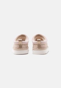 Ζευγάρι μπεζ παπούτσια slip-on με λευκές σόλες, ροζ και λευκές κεντητές λεπτομέρειες και πίσω θηλιές, σε λευκό απλό φόντο.