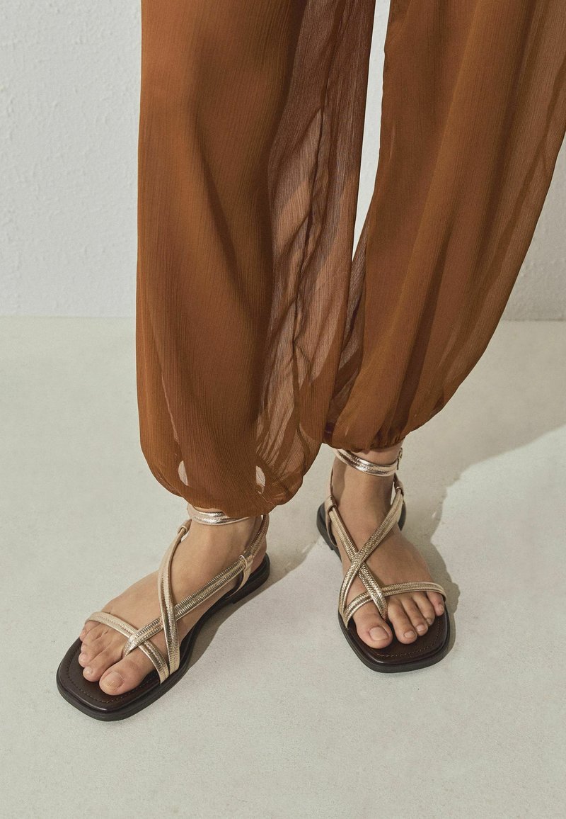 Pieds portant des sandales plates à brides métalliques dorées avec des bouts carrés et un pantalon marron transparent et ample, resserré aux chevilles.