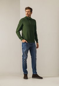Groen gebreide pullover met een halve rits kraag, gecombineerd met blauwe slim-fit jeans en bruine suède sneakers. Eenvoudig ontwerp, geen patronen of logo's.