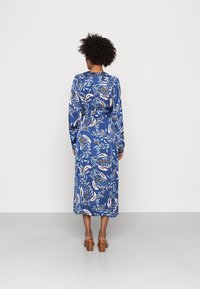 Frau mit lockigem Haar, die ein langärmliges blaues Midikleid mit Blumenmuster und braune Blockabsatzsandalen trägt und mit dem Gesicht zu einer weißen Wand steht.