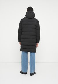 Doudoune noire avec capuche, présentant un matelassage horizontal. Portée sur un jean bleu clair ample et des bottes noires. Texture lisse, design mi-long.