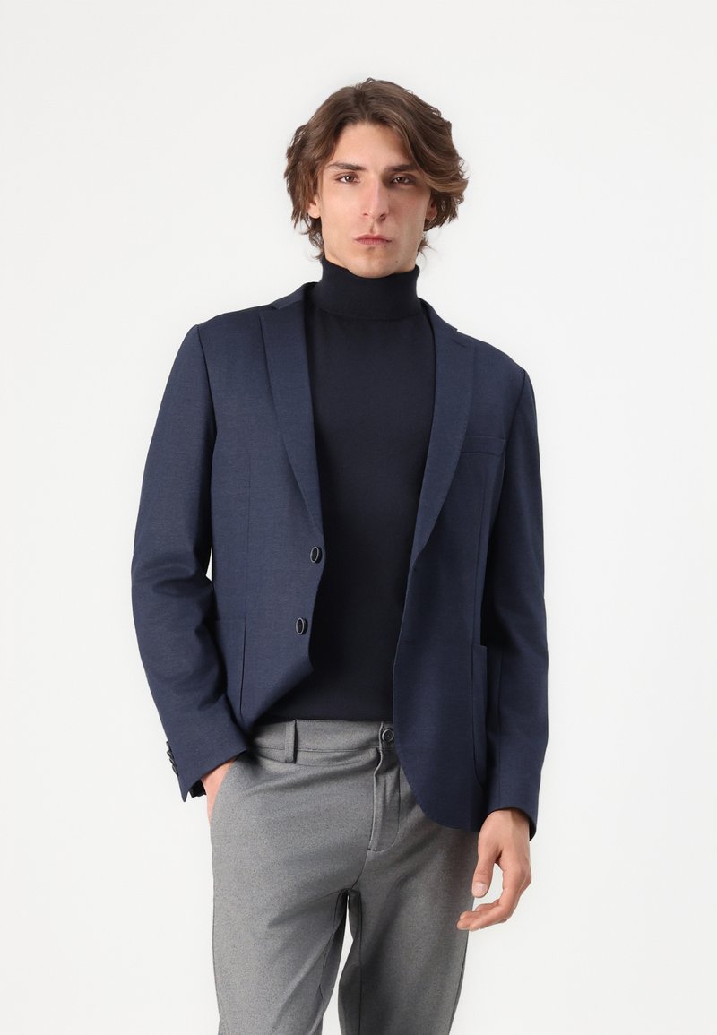 Blazer marine à deux boutons, coupe ajustée et revers en pointe. Porté sur un col roulé noir, accompagné de pantalons gris. Texture de tissu lisse.