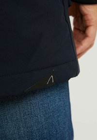 Granatowa kurtka z tkaniny o fakturowanym wykończeniu, z wyraźną czarną trójkątną metką w pobliżu dołu, zestawiona z dżinsami.