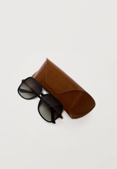 Lunettes de soleil carrées à monture noire avec verres dégradés reposant sur un étui à lunettes en cuir marron sur une surface blanche.