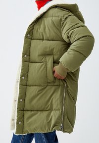 Abrigo acolchado de color verde oliva con capucha, con un forro de piel sintética, botones a presión, cremallera lateral y puños acanalados. Dos bolsillos frontales visibles.