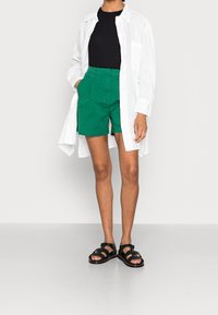 Biała koszula z długim rękawem, czarny dopasowany top, zielone szorty i czarne sandały. Koszula jest lekka; szorty mają boczne kieszenie, a sandały posiadają paski.