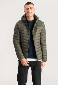 Olivengrøn puffjakke med quiltet struktur, lynlås lukning og hætte. Båret over en mørkeblå sweater, parret med sorte bukser.