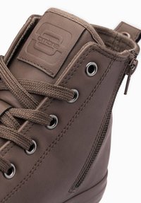 Bota de cuero marrón hasta el tobillo con punta redonda, superficie texturizada, ojales de metal y cierre lateral con cremallera. Presenta un parche de marca en la lengüeta.