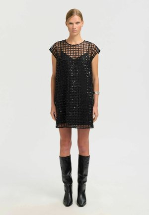 Woman wearing a black mesh sequin dress with a sleeveless black slip underneath, black knee-high boots, and a silver bangle on her left wrist.