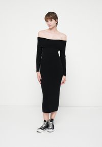 Rochie midi neagră, cu umeri descoperiți, cu mâneci lungi, design ajustat, realizată dintr-un material neted, asociată cu tenisi negri cu guler înalt.