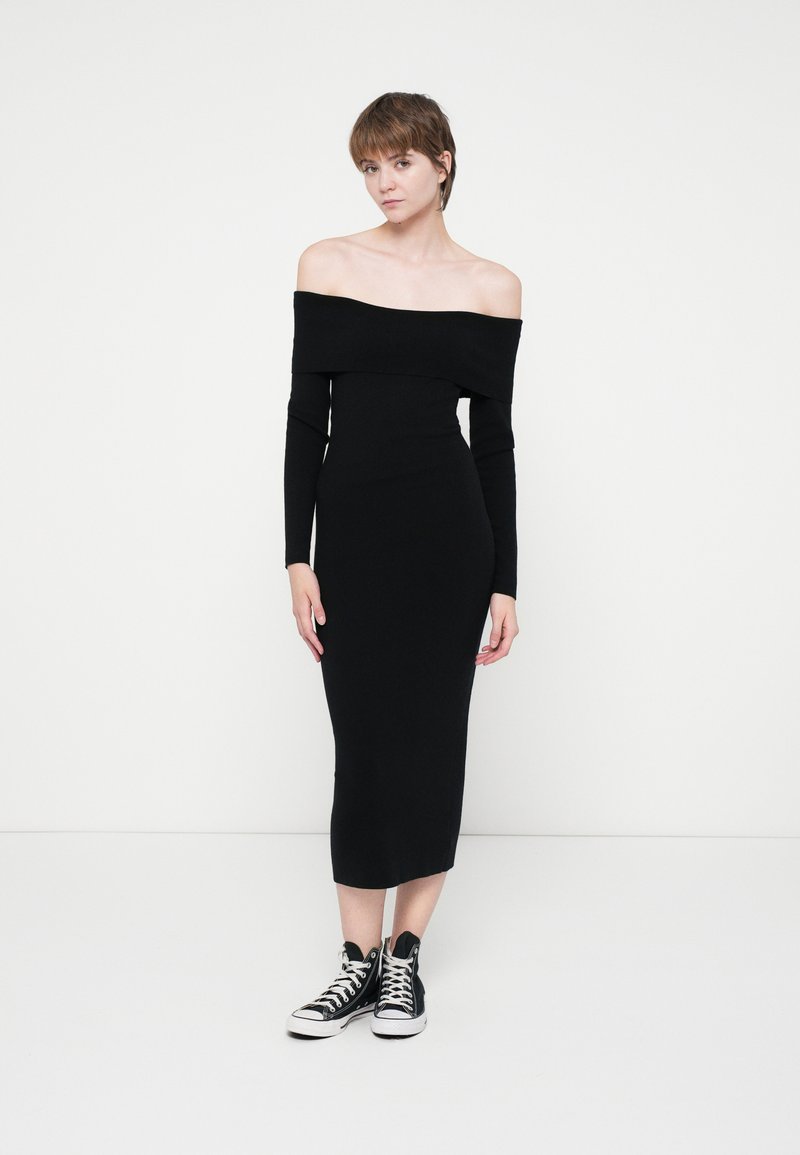 Rochie midi neagră, cu umeri descoperiți, cu mâneci lungi, design ajustat, realizată dintr-un material neted, asociată cu tenisi negri cu guler înalt.