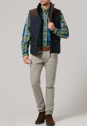 Gilet imbottito blu navy con accenti in pelle scamosciata marrone su una camicia a scacchi verde, blu e bianco. Pantaloni grigi chiari e stivali in pelle scamosciata marrone.