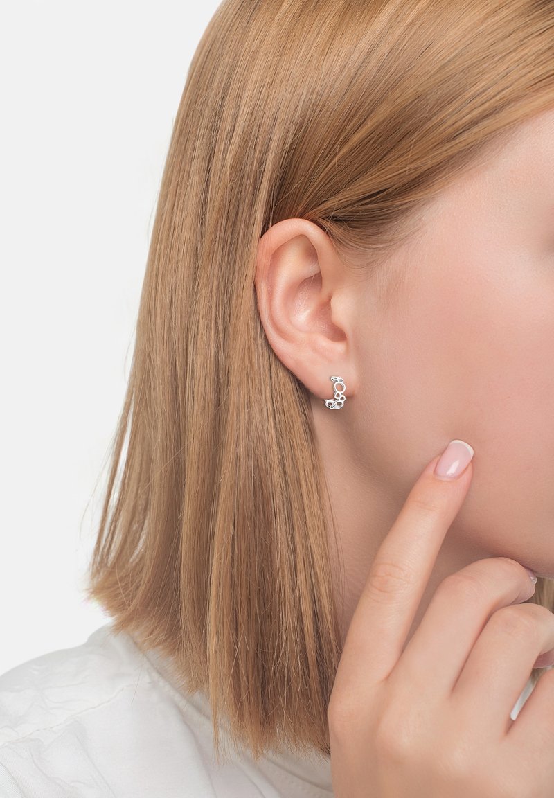 Gros plan d’une personne aux cheveux lisses châtain clair portant des boucles d’oreilles argentées en grappes circulaires, touchant sa joue avec un doigt manucuré.