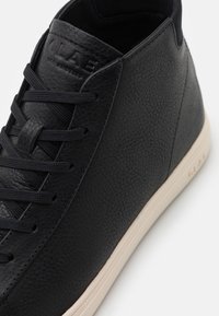 Černé kožené vysoké tenisky s texturovaným povrchem, s minimálním zavazováním a kontrastní béžovou gumovou podrážkou. Logo na jazyku.