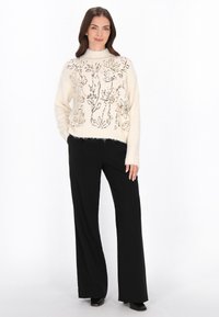 Kremowy sweter dzianinowy z kwiatowym haftem i cekinami, w połączeniu z czarnymi spodniami o szerokich nogawkach. Sweter ma prążkowany, wysoki dekolt.