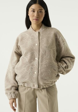 Frau trägt eine beige gemusterte Jacke mit Knöpfen und elastischen Bündchen sowie hochtaillierte beige Hose, steht vor einem schlichten Hintergrund.