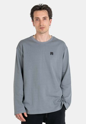 Langärmliges, graues T-Shirt aus Baumwolle mit einem runden Halsausschnitt und einem kleinen schwarzen Logo auf der linken Brust. Glatte Textur und lockere Passform.