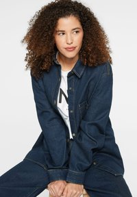 Denim jumpsuit in dark blue with a button-up front, chest pocket, and accent stitching. Worn over a white graphic t-shirt.