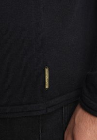 Pull bleu marine foncé en tissu tricoté texturé. Présente une petite étiquette rectangulaire dorée gravée de "SCOTCH". Ourlet simple.