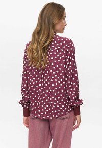 Femme avec de longs cheveux châtain clair portant un blouse long à manches maroon avec des pois blancs irréguliers et un pantalon rose ample, vue de dos.