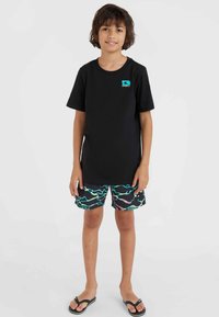 Camiseta negra de manga corta con logo turquesa, combinada con bañadores negros que tienen un patrón de olas coloridas. Usado con chanclas negras.