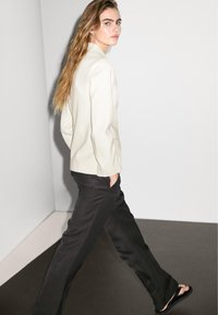 Frau mit langen Haaren geht drinnen und trägt ein weißes Blazer, schwarze Hose und schwarze Sandalen, während sie über ihre Schulter schaut.