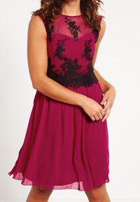 Žena ve vypasovaných magentových šatech s černou krajkovou aplikací stojí a jednou rukou drží sukni, čímž předvádí elegantní styl.