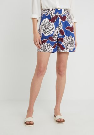 Frau trägt weißes Shirt und blaue Shorts mit großem weißem und rotem Blumenmuster, steht mit weißen offenen Sandalen auf einfarbigem Hintergrund.