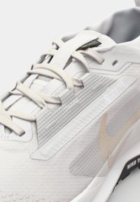 Lys grå sneaker med en tekstureret overdel, Gore-Tex mærke og tynde snørebånd. Har en diskret gylden swoosh og en gummisål.