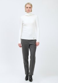 Vrouw staat in een witte ribbelige coltrui met ruches, grijze op maat gemaakte pantalons en zwarte puntige enkellaarsjes.