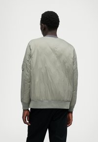 Cazadora bomber de color gris claro con una textura suave, puños y dobladillo acanalados, y costuras decorativas en las mangas. Posee un clásico cuello redondo.